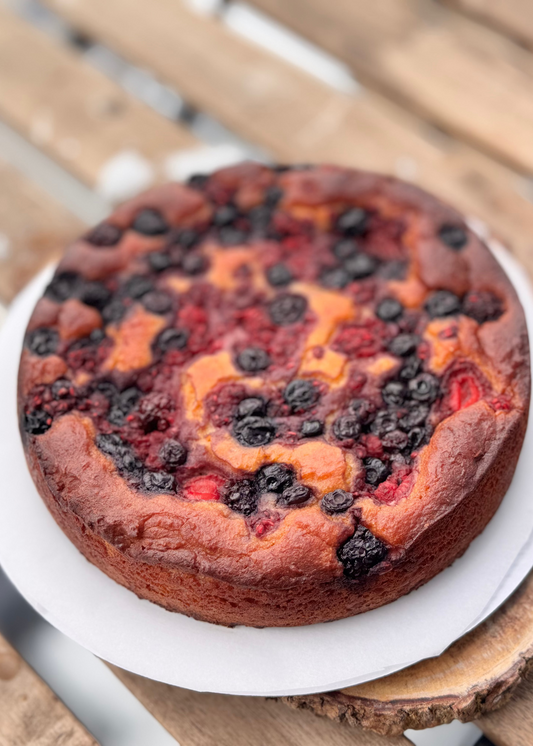 Round sugar-free, keto, gluten-free berry-topped cake on a white parchment-lined plate, featuring baked mixed berries and golden cottage-cheese batter swirled across the surface, set on a rustic wooden table.