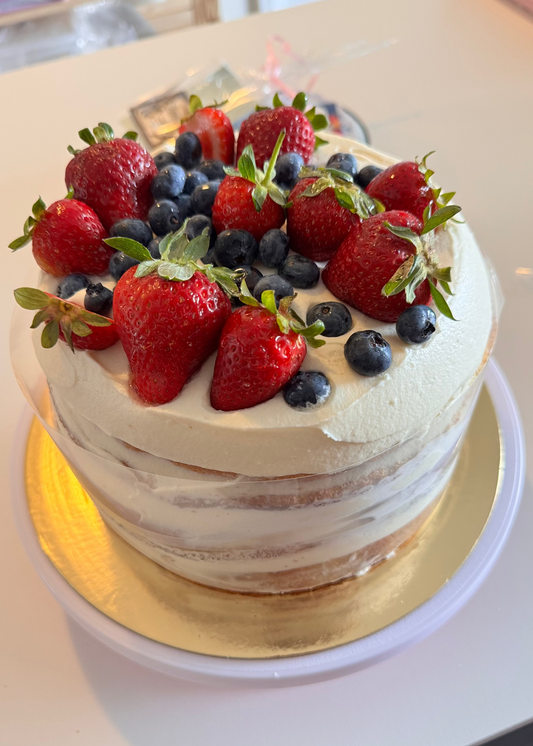 A semi-naked cake with white frosting, topped with fresh strawberries and blueberries, sitting on a gold cake board on a white table.