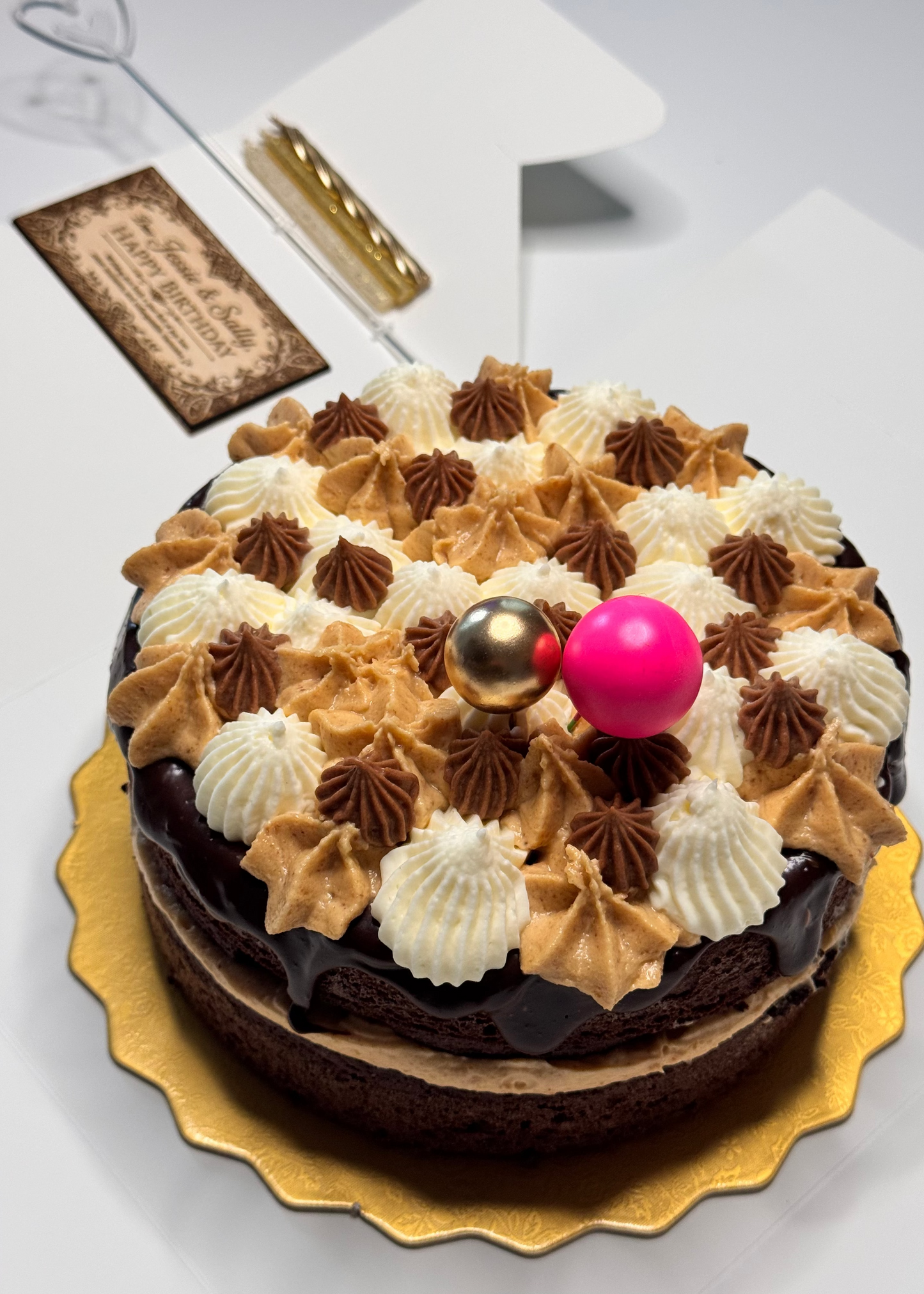 A chocolate cake decorated with piped cream and brown frosting swirls, topped with two gold spheres and a bright pink ball, served on a gold scalloped plate against a white background.