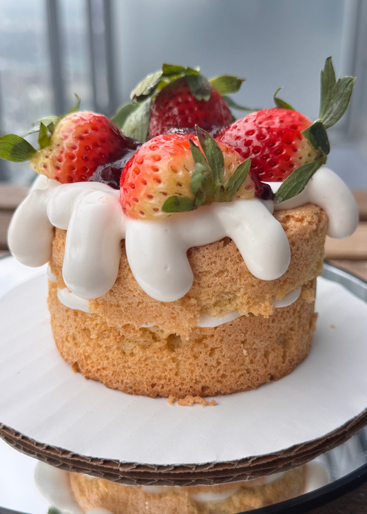 4" layered sponge cake topped with fresh strawberries and glossy marshmallow frosting, sitting on a white cake board with soft natural light and a blurred window background.